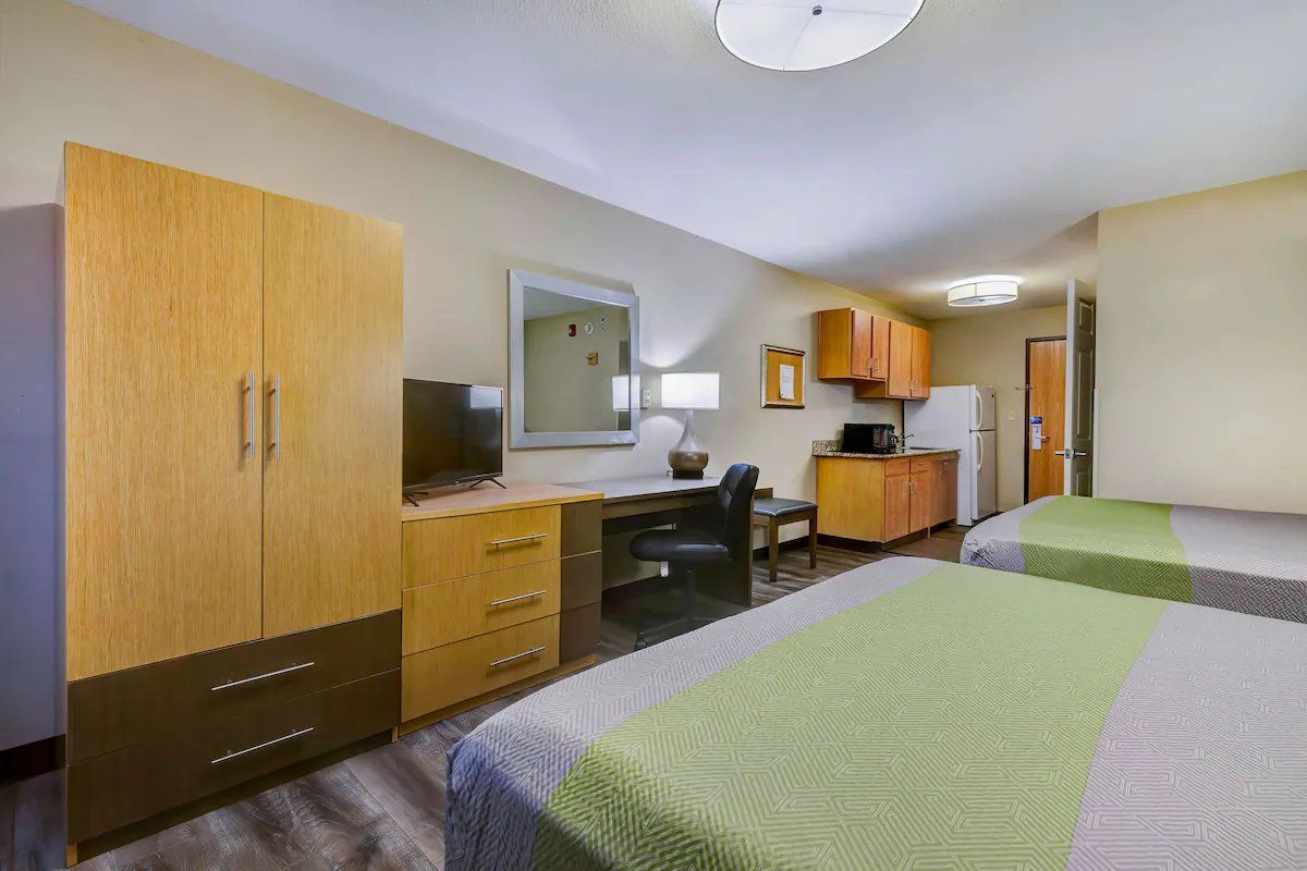 A hotel room with two beds, a desk, a chair, a television, and a kitchenette.