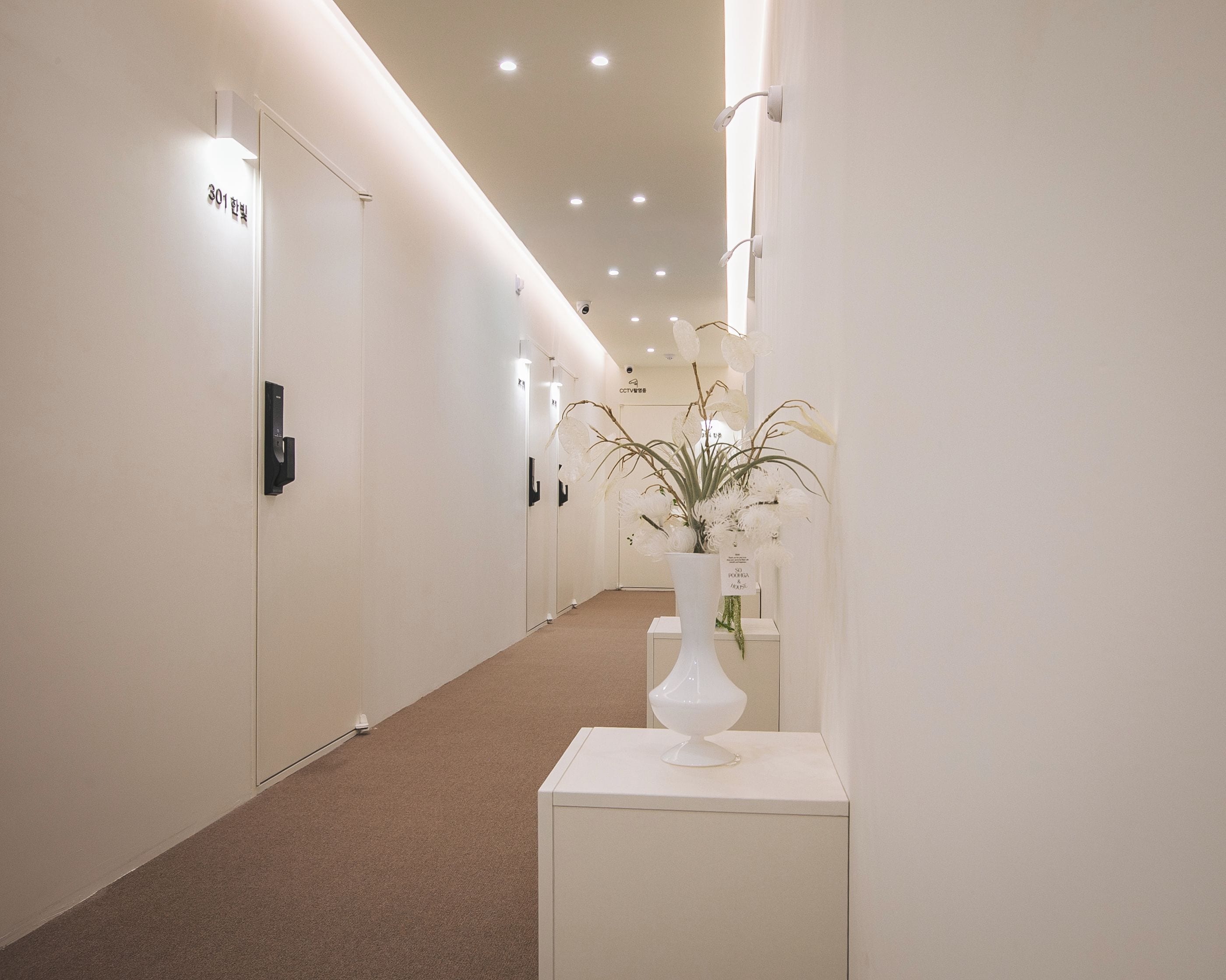 A hallway with white walls, doors, and a carpeted floor. There is a white vase with flowers on a pedestal.