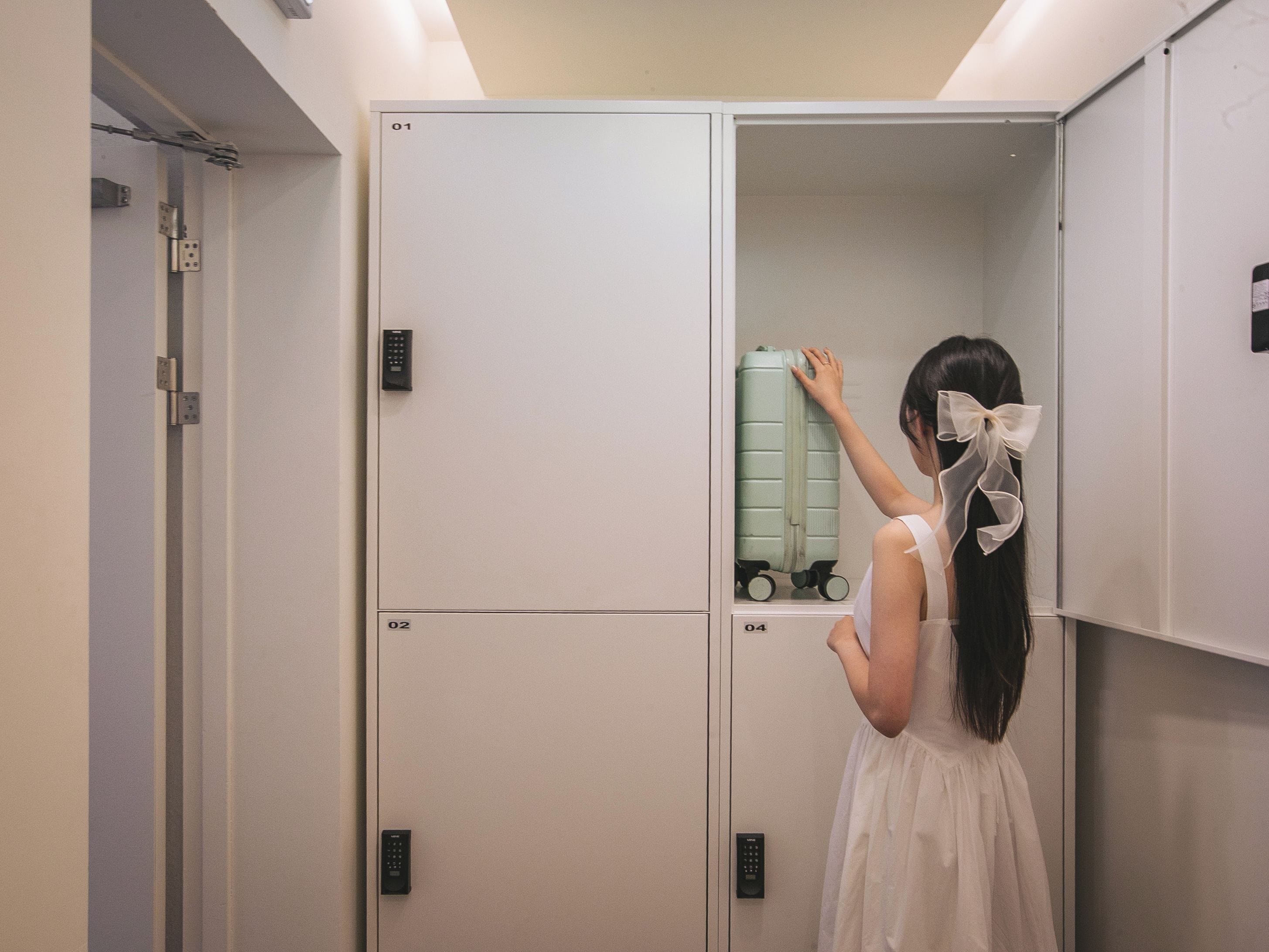 A person in a white dress with a bow in their hair, standing in front of a set of lockers, with a suitcase on one of the lockers.