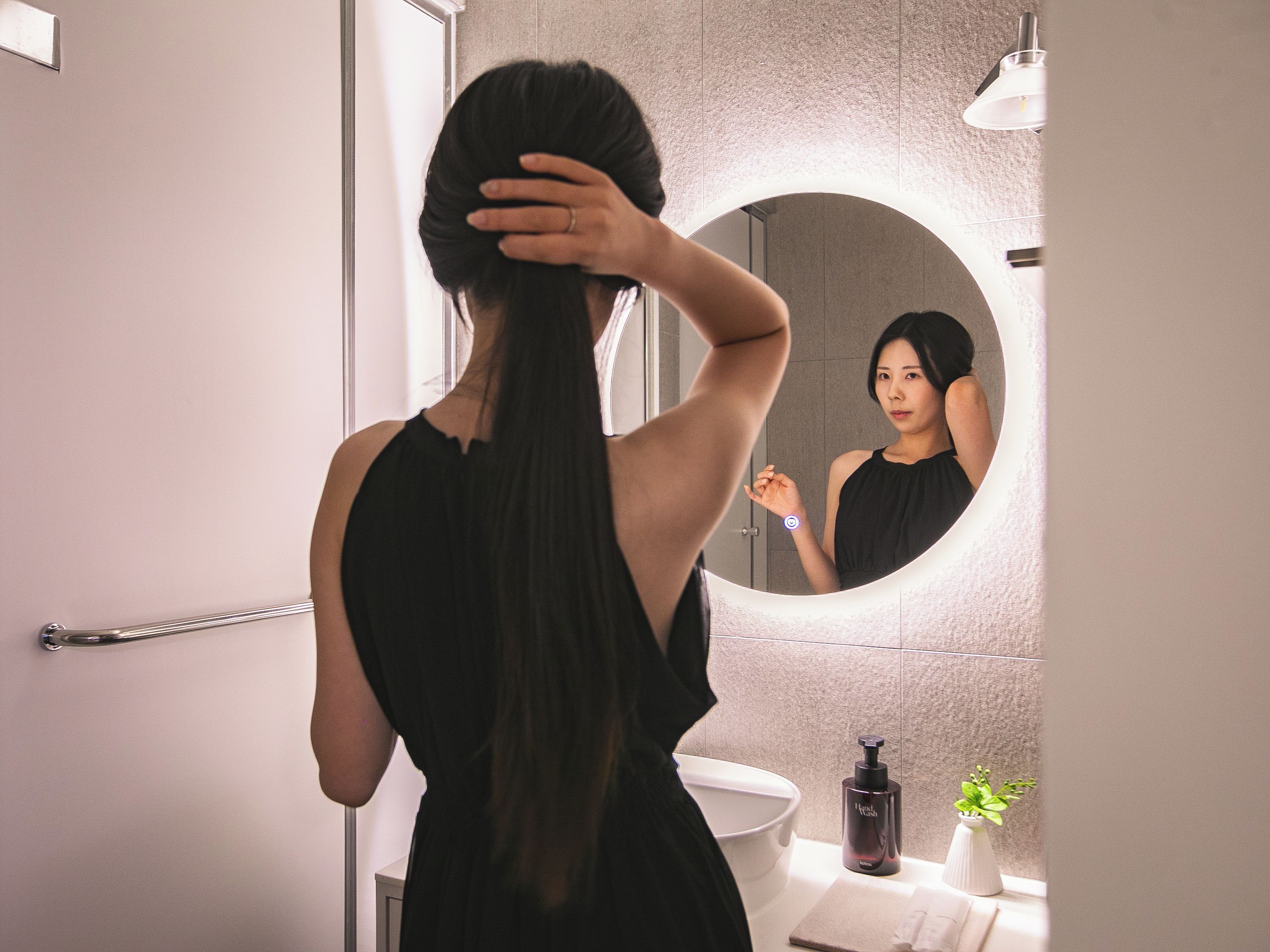 A person with long hair in a bathroom with a round mirror, a sink, and a bottle of hand soap.