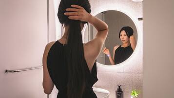 A person with long hair in a bathroom with a round mirror, a sink, and a bottle of hand soap.