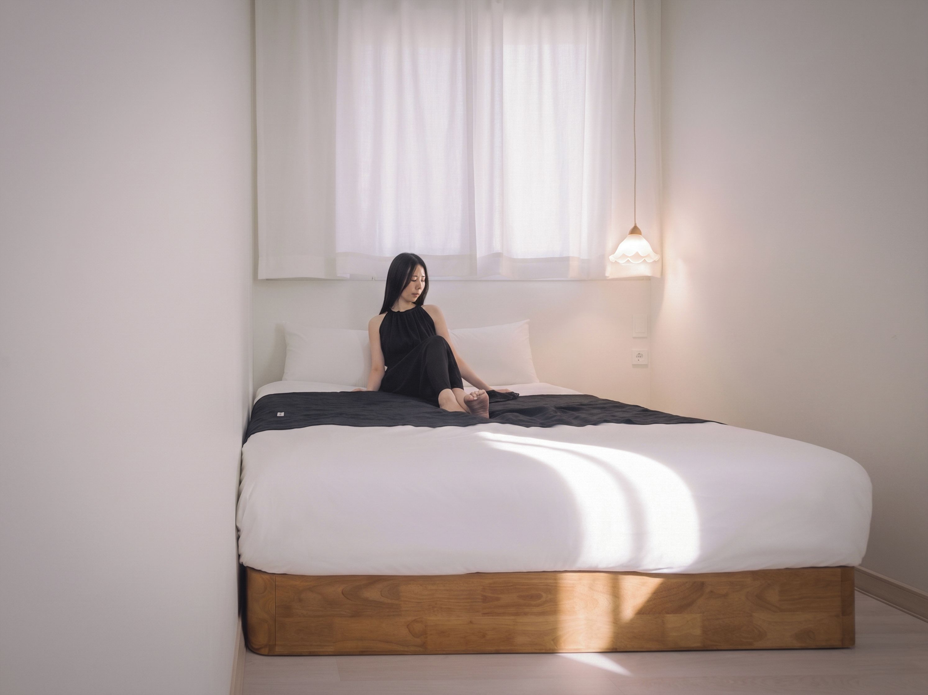 Una cama con estructura de madera y ropa de cama blanca, una persona sentada en la cama y una lámpara de noche.