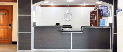Una reception con una scrivania, uno schermo digitale, un cartello con il numero '6' e una telecamera di sicurezza.