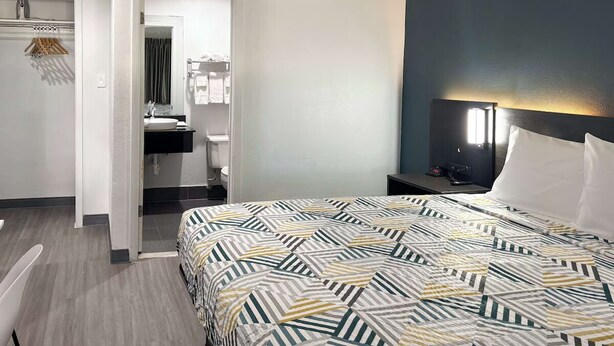 Ein Hotelzimmer mit Bett, Nachttisch, Lampe, Spiegel, Badezimmer mit Waschbecken und Handtuchhalter sowie einer Tür zu einem weiteren Raum.