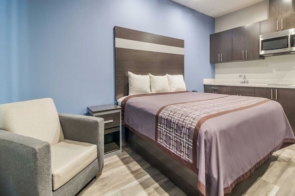 A hotel room with a bed, a nightstand, a chair, a kitchenette, and a blue wall. - Extended Stay Near Houston Industrial Area – Within 7 Miles (Deer Park)