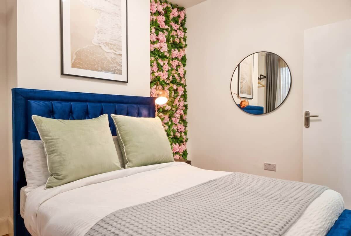 A bedroom with a blue headboard, a bed with white and green bedding, a wall-mounted mirror, and a floral wall decoration.