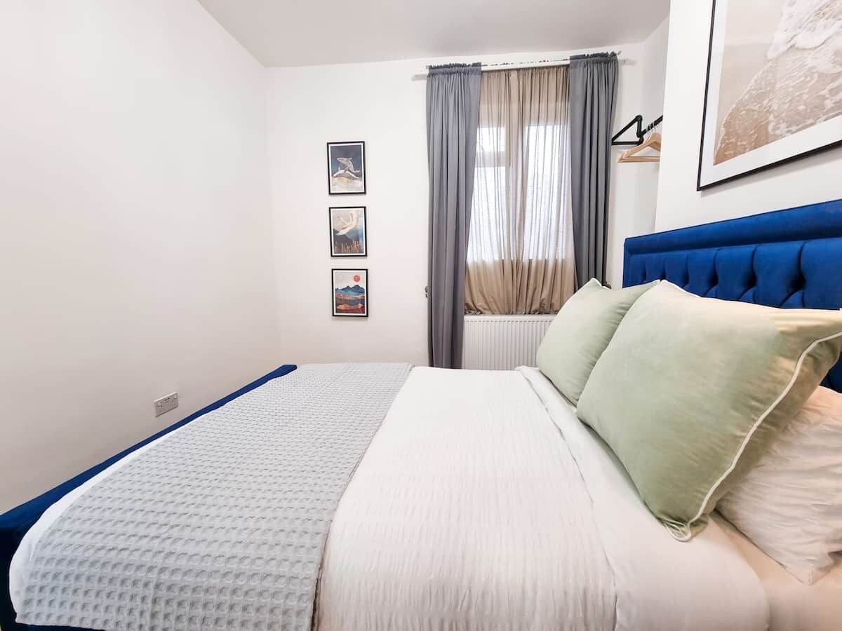 A bedroom with a bed, a blue headboard, a window with curtains, and three framed pictures on the wall.