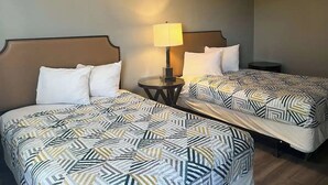 A hotel room with two beds, each with a patterned bedspread, a bedside table with a lamp, and a small round table.