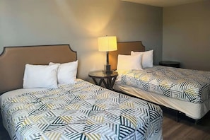 A hotel room with two beds, each with a patterned bedspread, a bedside table with a lamp, and a small round table.