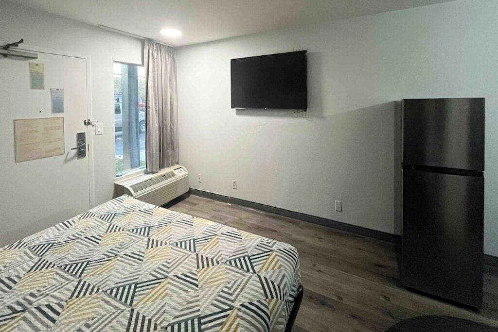 A hotel room with a bed, a flat-screen TV mounted on the wall, a built-in air conditioning unit, and a refrigerator.