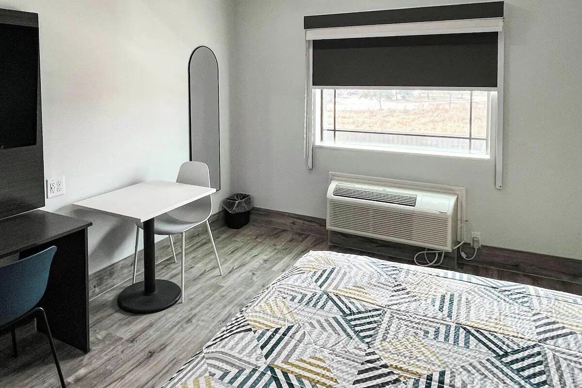 Una habitación con cama, escritorio, silla, lámpara de pie, ventana con vistas y radiador.