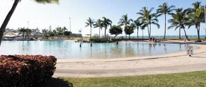 A large swimming pool with a sandy beach area, surrounded by palm trees and a clear sky.