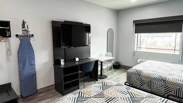 Habitación de hotel con cama, escritorio y silla, televisor, tabla de planchar y ventana.