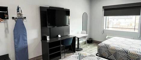 Una camera d'albergo con un letto, una scrivania con sedia, una TV, un asse da stiro e una finestra.