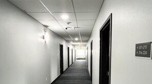 A long, narrow hallway with doors on both sides, a carpeted floor, and a ceiling with recessed lighting and smoke detectors.