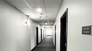 A long, narrow hallway with doors on both sides, a carpeted floor, and a ceiling with recessed lighting and smoke detectors.
