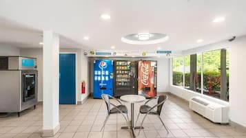 A clean, well-lit indoor area with vending machines, a table with chairs, and large windows.