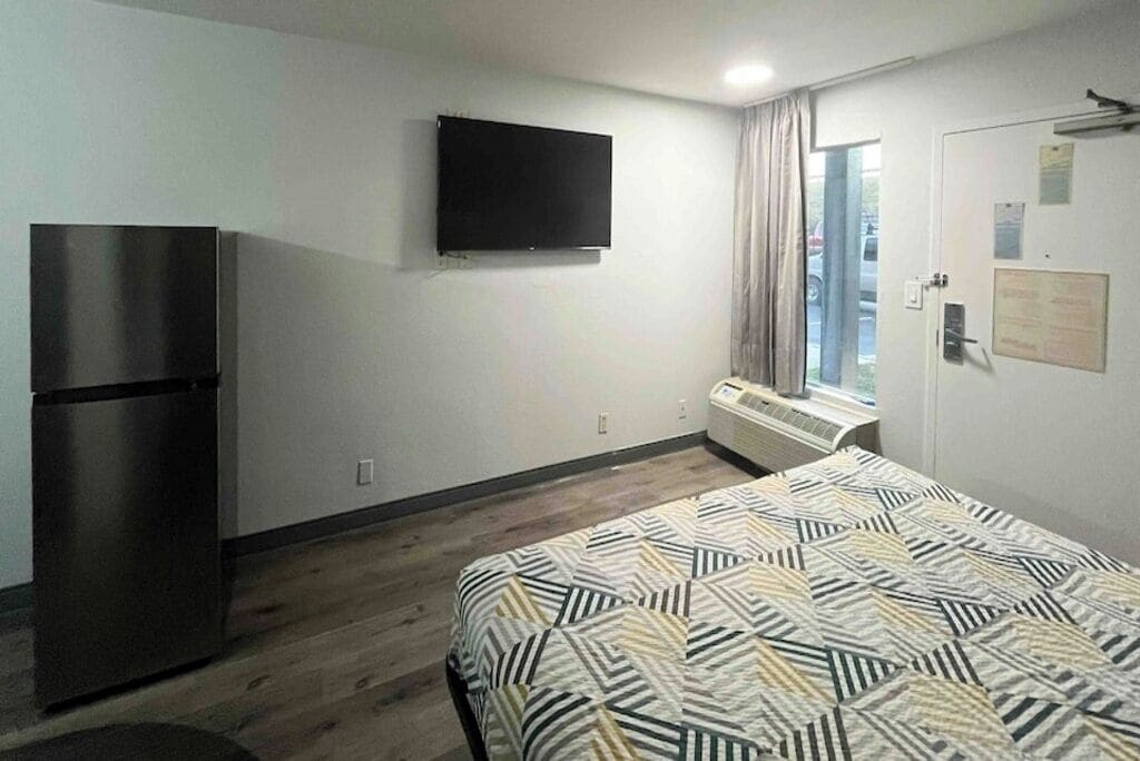 A hotel room with a bed, a flat-screen TV, a refrigerator, and a window with curtains.