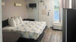 A hotel room with a bed, bedside table, a small desk, a chair, a window with curtains, and a door with a notice. - Augusta King Room w/ Kitchenette Near Riverwalk Entertainment (Augusta)