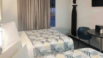 A hotel room with a bed, a chair, a table, a window with curtains, and a wall-mounted item.