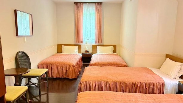Una habitación de hotel con dos camas individuales, una mesita pequeña con una silla, una mesita de noche y una ventana con cortinas.