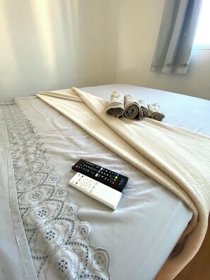 Una cama con una manta blanca, una toalla enrollada y un control remoto.