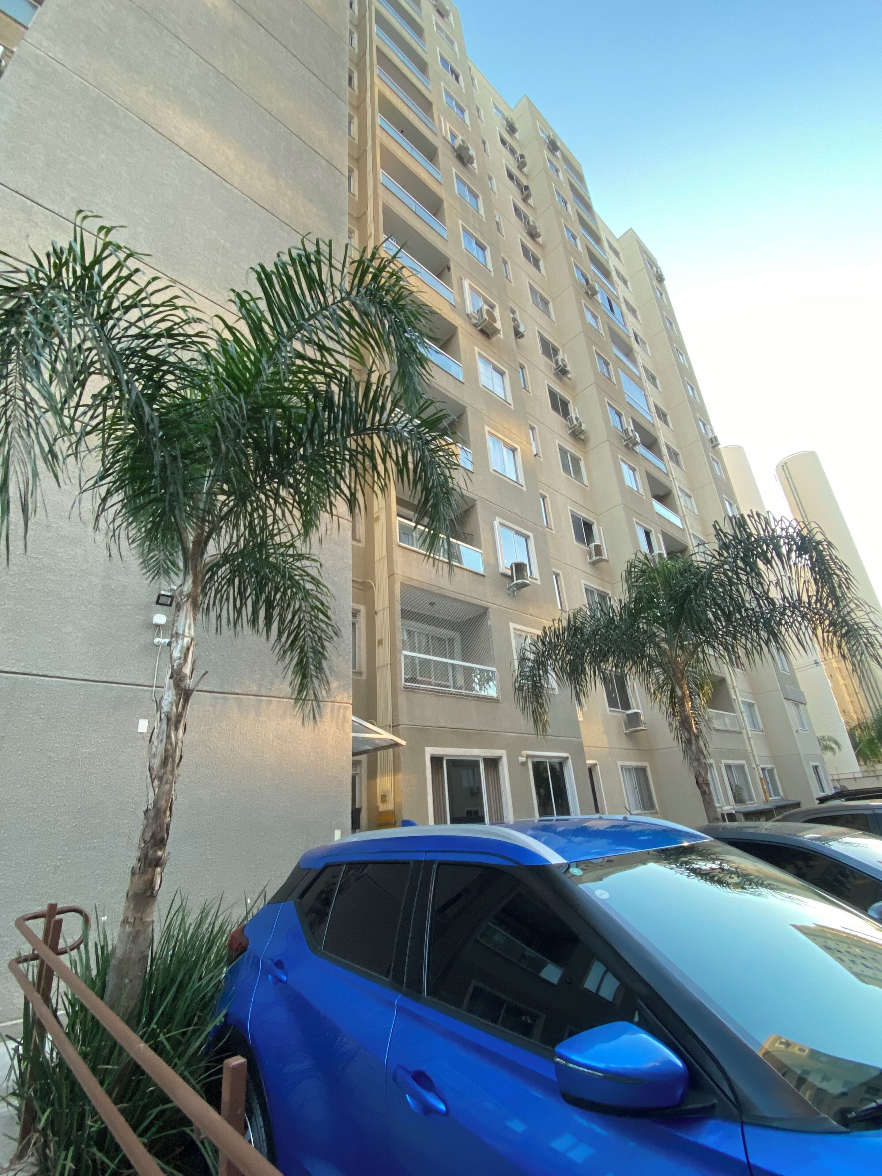 A multi-story residential building with balconies, a parked blue car, and palm trees.