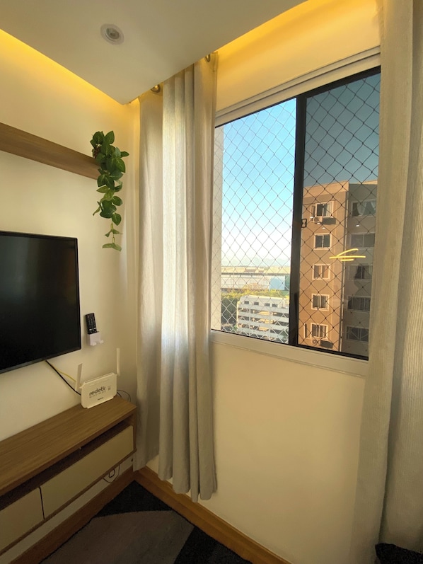 A room with a TV, a redbud router, a plant, and a window with a view of buildings. - Apartment near Galeão Airport • 2 bedrooms • With 1 vacancy (Duque de Caxias)