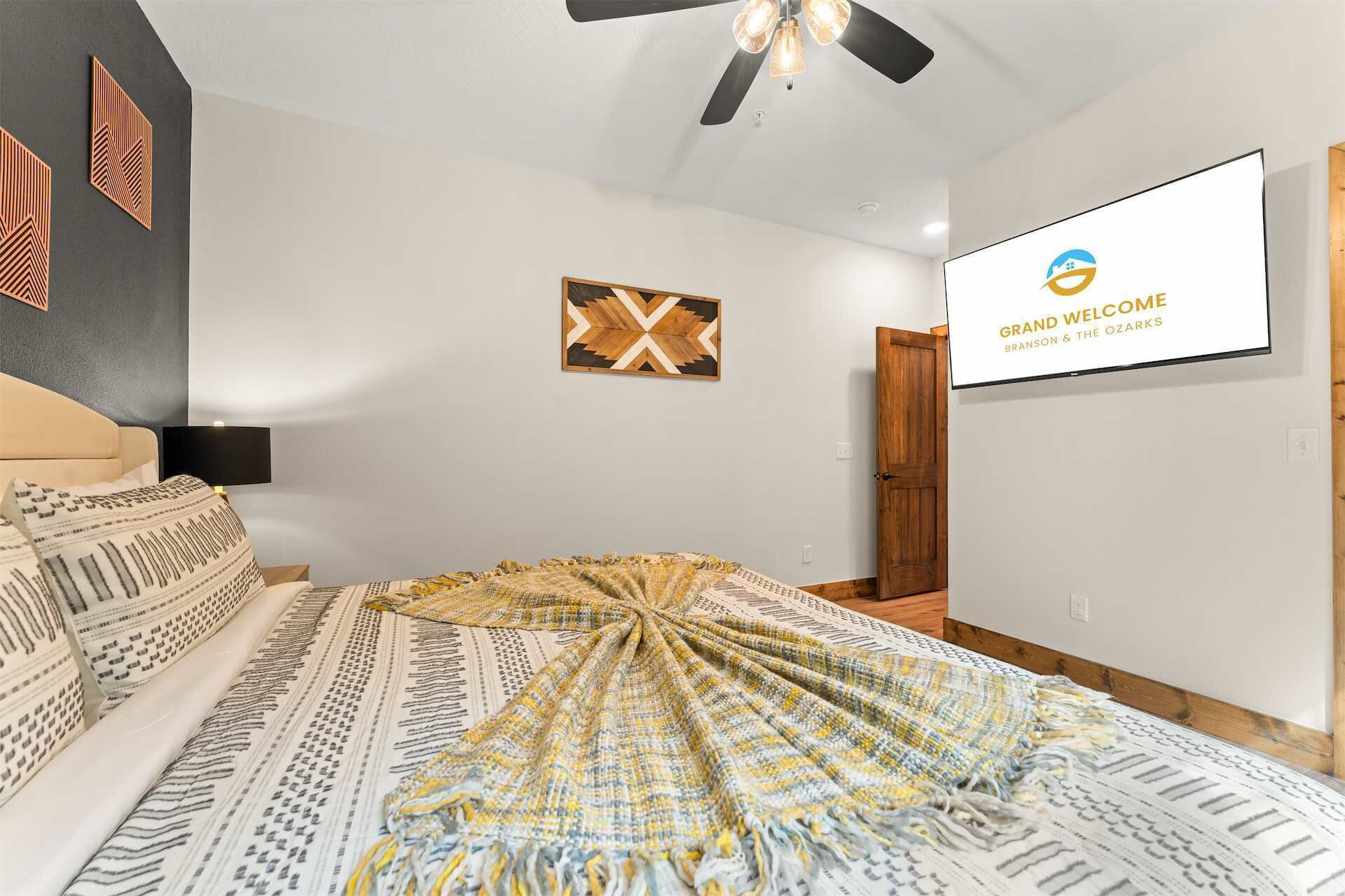 Ein modernes Schlafzimmer mit einem Bett, einem Deckenventilator, einem an der Wand montierten Fernseher und dekorativer Wandgestaltung.
