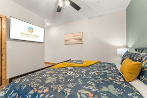 A bedroom with a bed, a ceiling fan, a wall-mounted TV displaying 'GRAND WELCOME BRANSON & THE OZARKS', and a framed artwork. - Creekside Cabin Cozy Stay (Hollister)