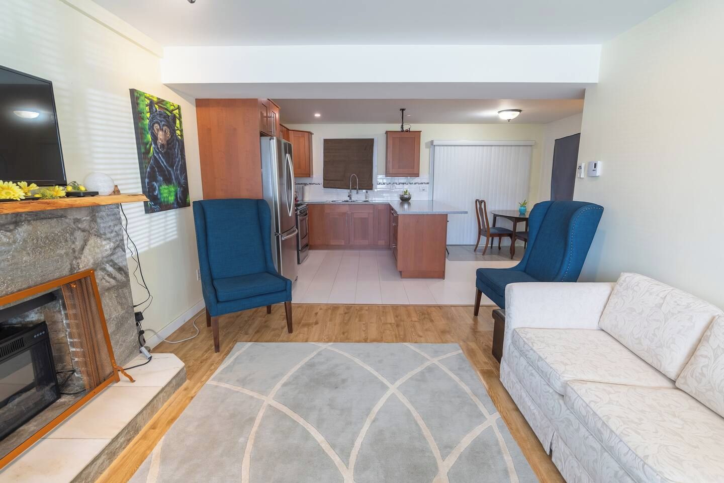 Un salon avec une cheminée, deux fauteuils bleus, un canapé blanc et un coin cuisine doté de meubles en bois et d’une table à manger.