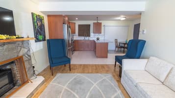 Un salon avec une cheminée, deux fauteuils bleus, un canapé blanc et un coin cuisine doté de meubles en bois et d’une table à manger.