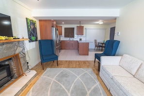 A living room with a fireplace, two blue armchairs, a white sofa, and a kitchen area with wooden cabinets and a dining table.