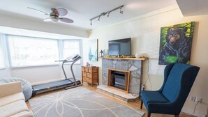 A living room with a treadmill, a fireplace, a blue armchair, and a bear painting on the wall. - Langley Suite Chef Kitchen (Langley Township)