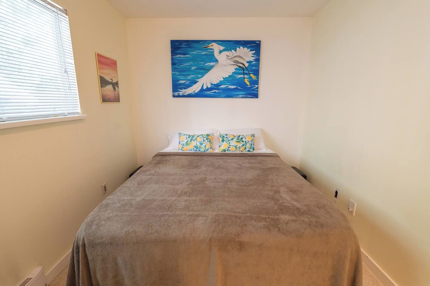 Une chambre à coucher avec un lit, une fenêtre avec des stores, un tableau représentant un oiseau et un interrupteur fixé au mur.