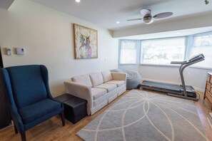 A living room with a sofa, a blue armchair, a treadmill, and a large window with blinds.
