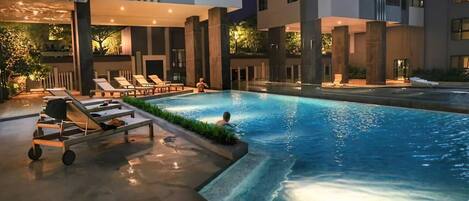 Una piscina exterior con tumbonas y un carrito junto a la piscina, rodeada de edificios modernos.