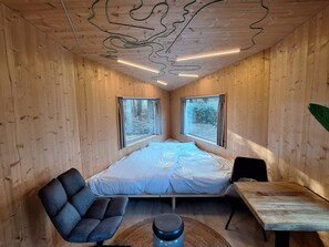 Quarto compacto com cama, cadeira, mesa e amplas janelas.