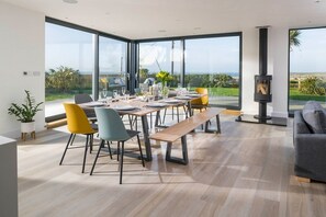 A modern dining area with a large table set for six, surrounded by large windows offering a view of the outdoors. - Dune House - sleeps 8 guests  in 4 bedrooms (Hayle)