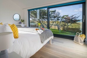 A modern bedroom with a large window, a bed with white bedding, a bedside table, and a view of trees. - Dune House - sleeps 8 guests  in 4 bedrooms (Hayle)