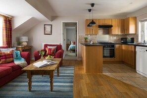 A modern kitchen with wooden cabinets, a central island, and a dining area with a red sofa and a coffee table. - Jasmine Cottage - sleeps 3 guests  in 2 bedrooms (Port Gaverne)