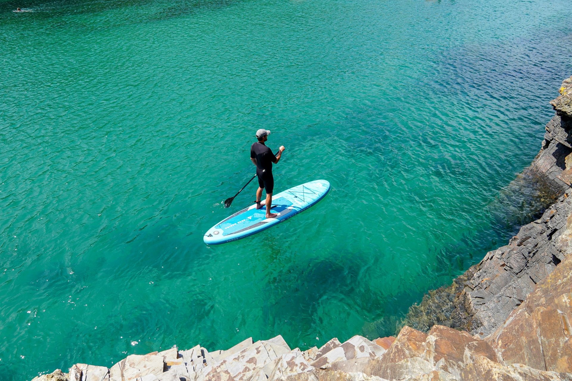A person paddleboarding on clear turquoise water near a rocky shoreline.