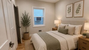 Ein Schlafzimmer mit Bett, Nachttischen, einem Fenster, einer Pflanze und einer Tür.