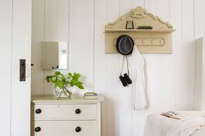Ein Zimmer mit einem weißen Schminktisch, einem Spiegel, einem Regal mit Hut und Fernglas sowie einem Bett mit weißer Decke.