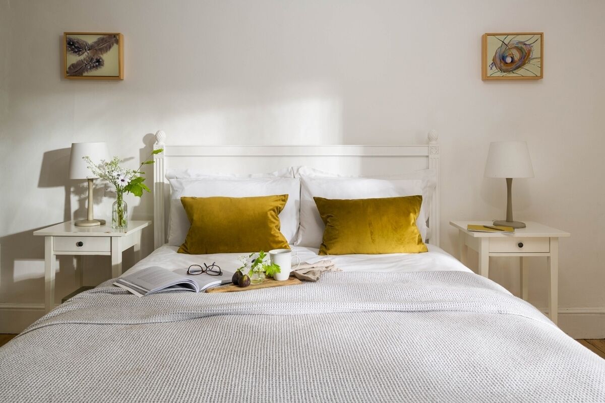 A neatly made bed with white bedding and two mustard pillows, flanked by white nightstands with lamps and a small table with a book and a cup.