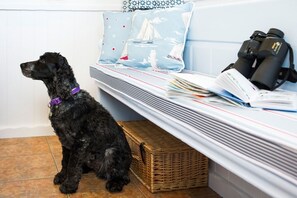 A black dog sitting on a tiled floor next to a bed with a blue pillow, a wicker basket, and a pair of binoculars on the bed. - Lighthouse View - sleeps 4 guests  in 2 bedrooms (Hayle)