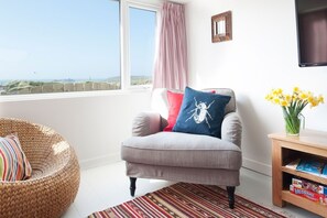 A room with a grey armchair, a wicker chair, a wooden side table with a vase of flowers, and a view of the sea through a window. - Lighthouse View - sleeps 4 guests  in 2 bedrooms (Hayle)