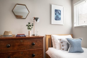 Ein Schlafzimmer mit einem hölzernen Bett, einer Kommode mit Schubladen, einem Spiegel, einer Lampe, einem gerahmten Bild und einem Fenster mit Jalousien.