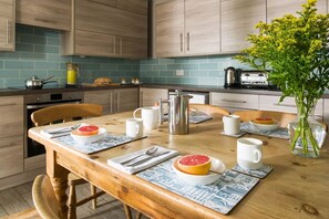 A kitchen with wooden cabinets, a dining table set with plates, cups, and a vase with flowers. - Lower Tregudda Apartment - sleeps 8 guests  in 5 bedrooms (Port Gaverne)