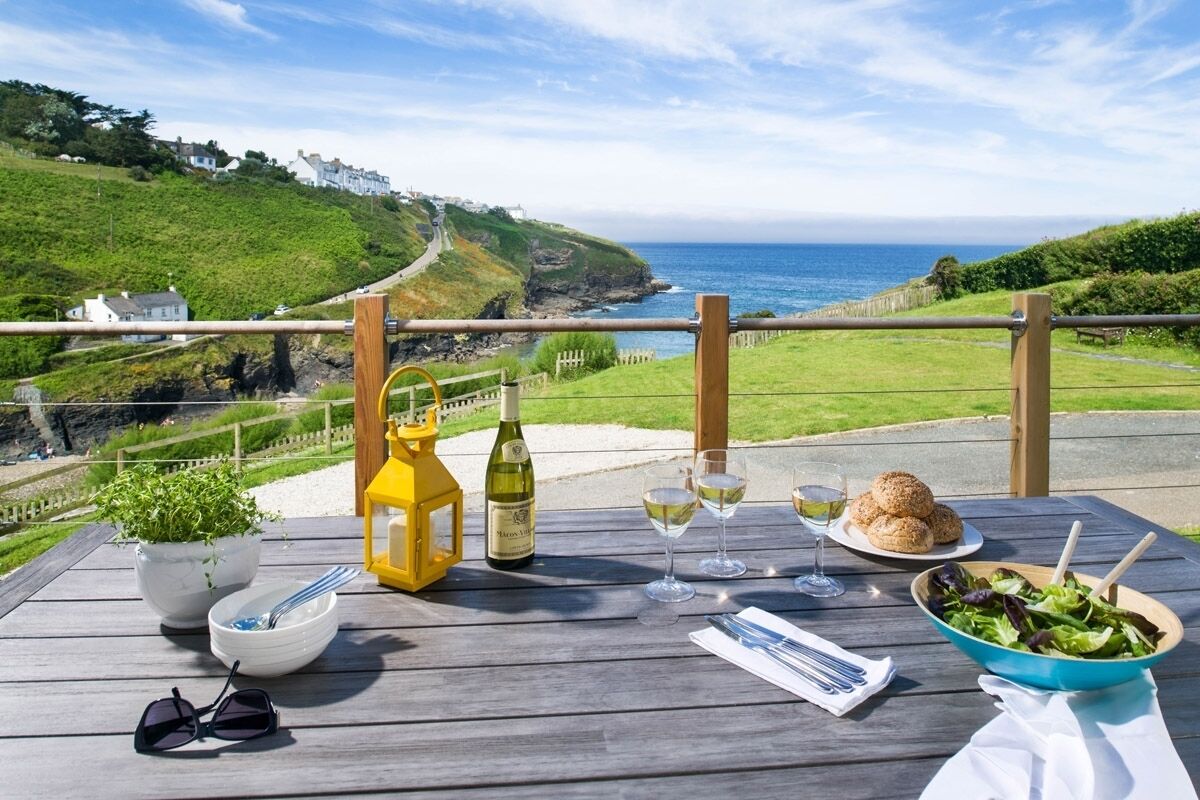 A table set outdoors with a view of the sea, a bottle of wine, glasses, a bowl of salad, and a plate of bread.
